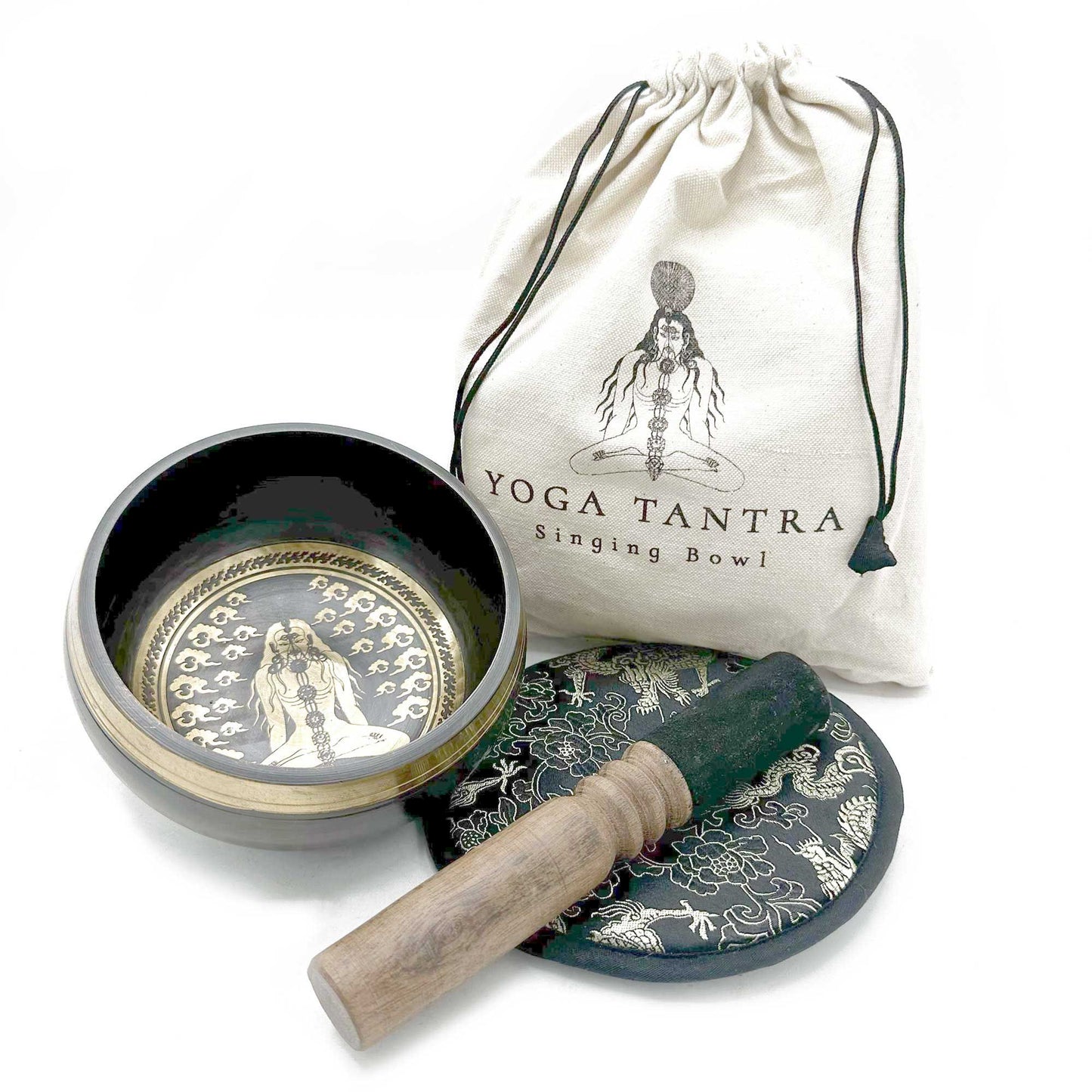 Singing bowl sits open revealing an etched meditating figure, resting beside a wooden mallet and embroidered cushion in front of a white drawstring bag labeled YOGA TANTRA Singing Bowl on a white background.