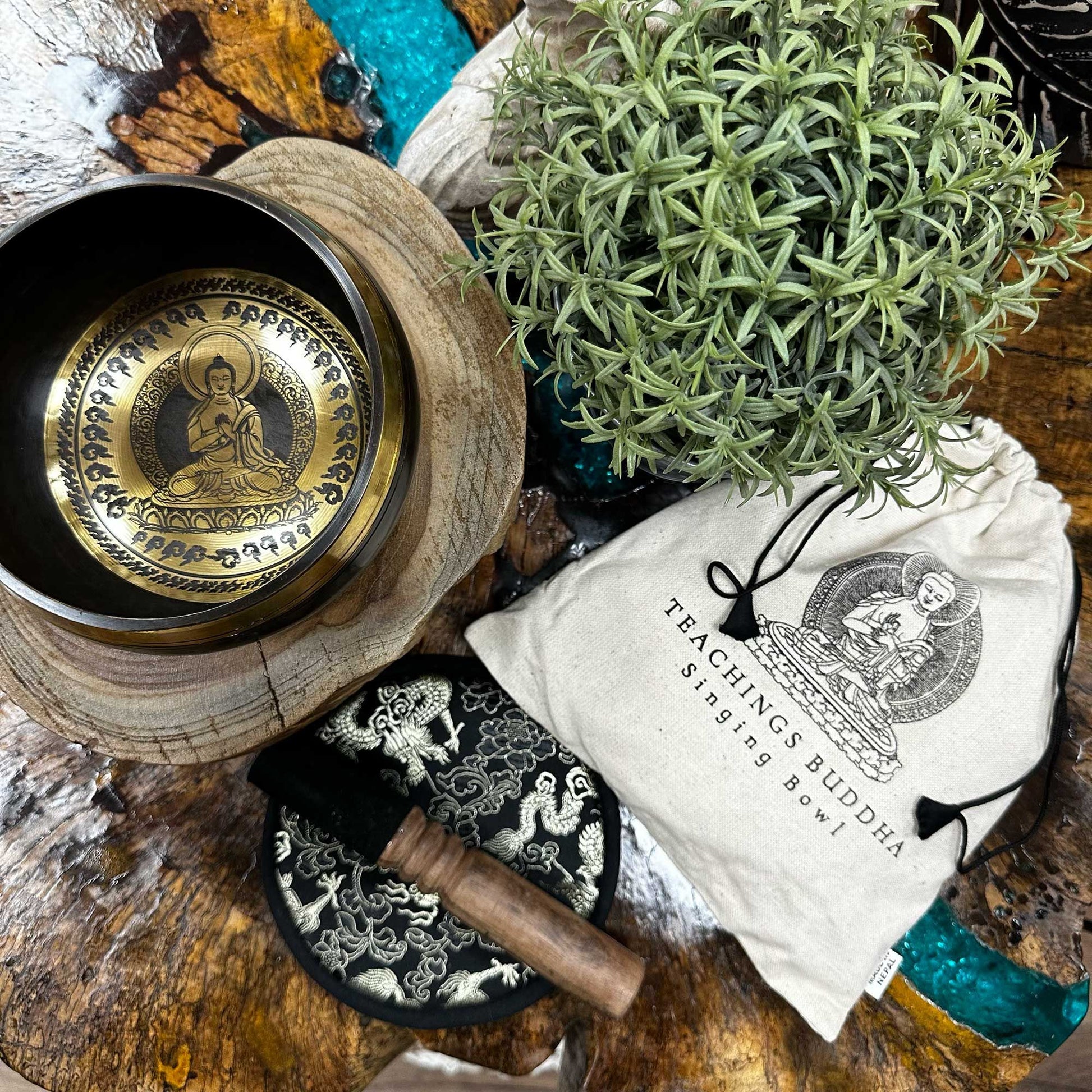 Brass singing bowl featuring an engraved Buddha and mantra "OM MANI PADME HUM" rests on a wooden slab beside a mallet, patterned cushion, a pouch labeled "TEACHINGS BUDDHA Singing Bowl" and a potted succulent; tag "NEPAL".