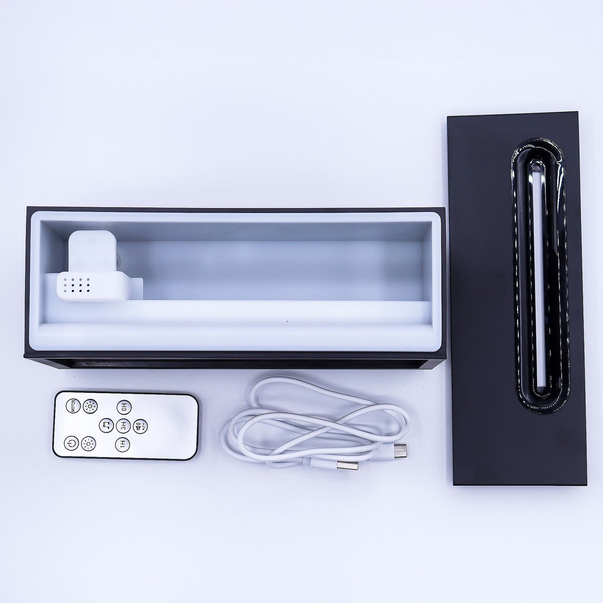 Rectangular tabletop diffuser sits disassembled with its black lid removed, inner white reservoir exposed, remote control and coiled USB cable arranged beside it on a plain white surface.