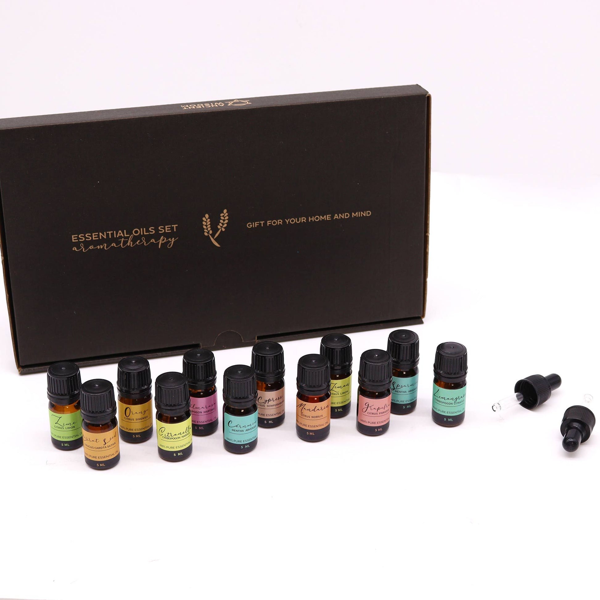 A row of small amber essential oil bottles arranged in front of a black gift box on a white surface; box text reads ESSENTIAL OILS SET aromatherapy and GIFT FOR YOUR HOME AND MIND; bottles marked 5 ML.