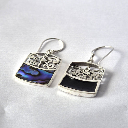 Pair of rectangular silver earrings display filigree tops and abalone blue inlay on one and black enamel on the other, resting on white paper backdrop, stamped '925' on one piece.