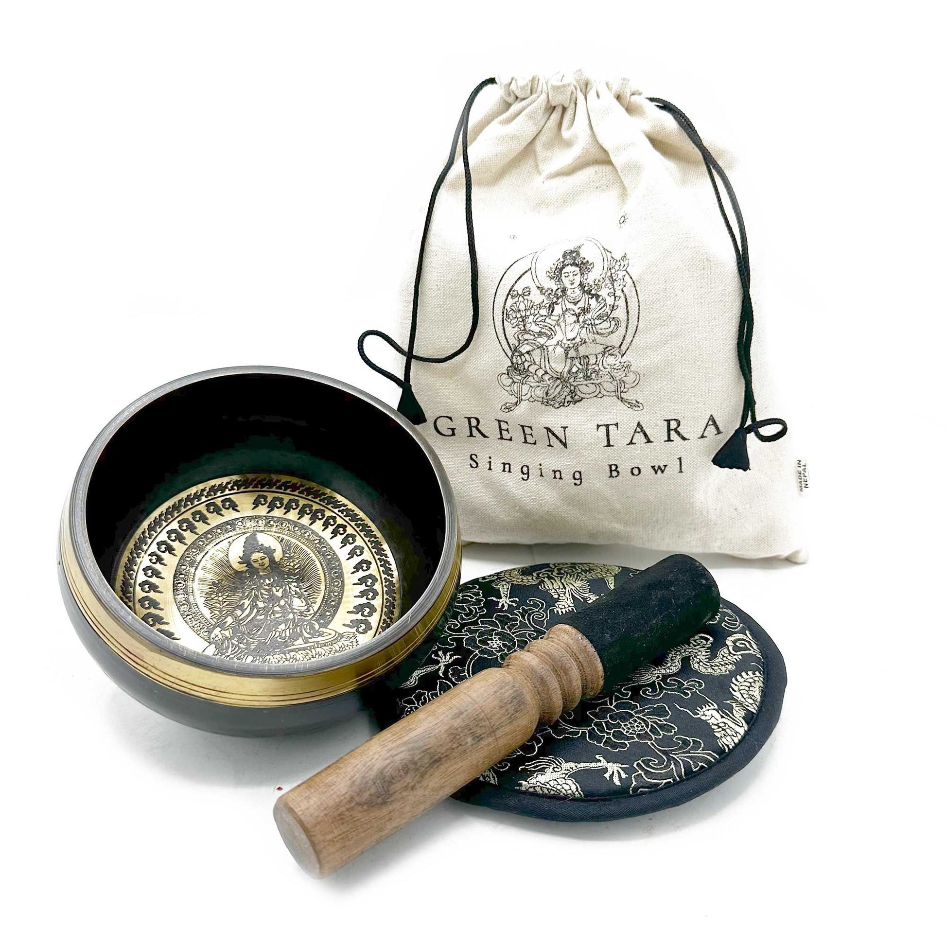 Brass singing bowl sits open with wooden mallet resting on an embroidered cushion and a drawstring cloth bag behind on a white surface; text on bag: GREEN TARA Singing Bowl.