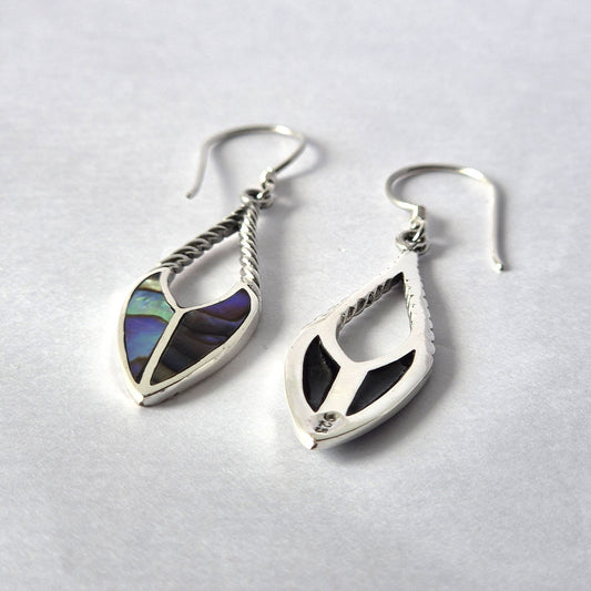 Silver teardrop earrings with twisted stems, one showing iridescent abalone inlay and the other black inlay stamped 925, lying on a white paper surface in close-up.