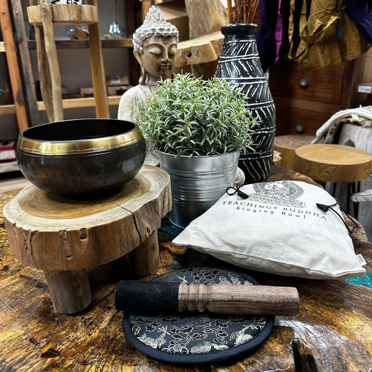 Singing bowl rests on a small wooden stool beside a mallet, potted plant and a fabric pouch labeled TEACHINGS BUDDHA Singing Bowl on a rustic wooden table with a Buddha statue in the background.