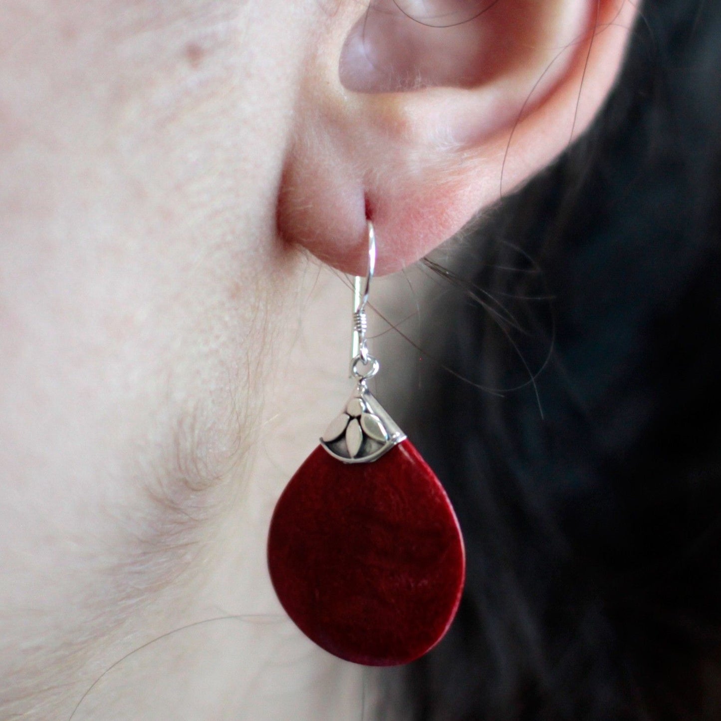 A red teardrop earring hangs from a pierced earlobe, suspended by a silver hook and decorative cap against pale skin with fine hairs and a dark blurred background.