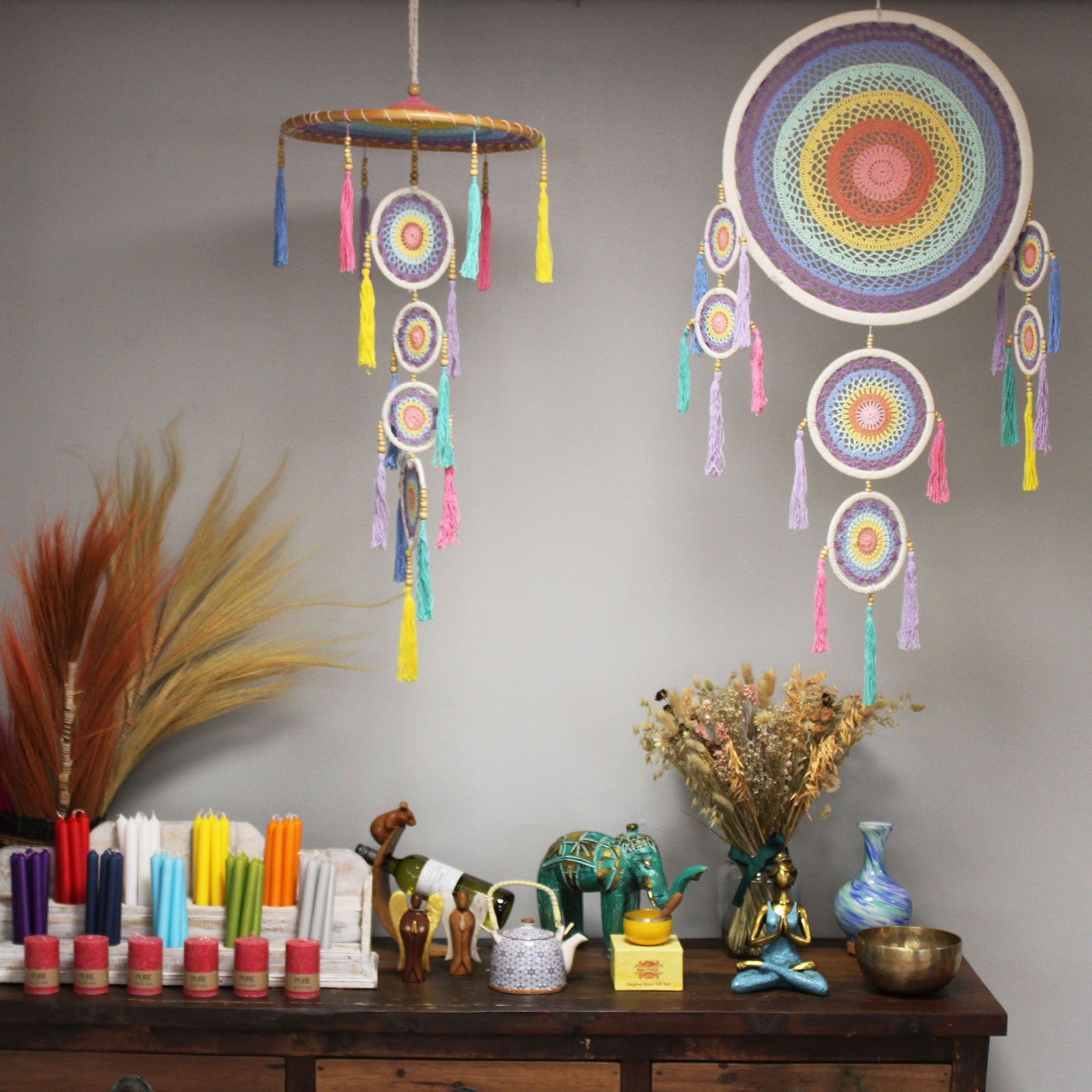 Colorful crocheted dreamcatchers hang above a wooden sideboard; the sideboard holds dried flowers, candles, a teapot, small figurines, an elephant and decorative vases against a gray wall.