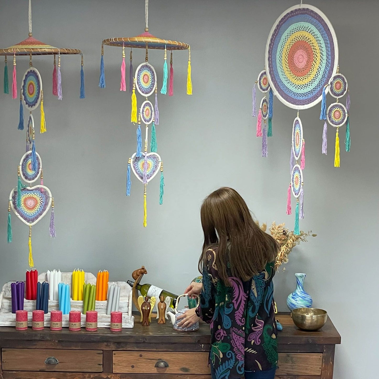 A woman pours from a teapot on a wooden sideboard while colorful crocheted dreamcatchers hang overhead in a shop; rows of candles and jars labeled PURE sit on the counter.