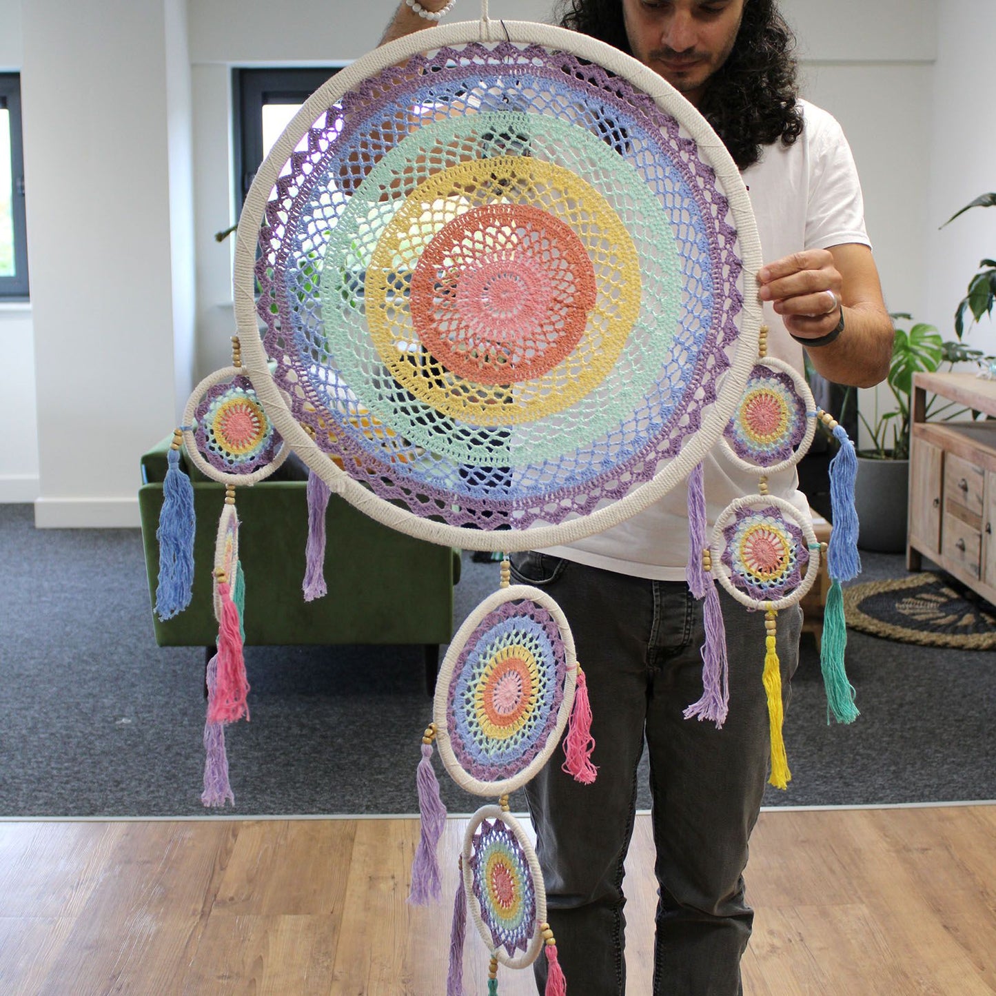 A large multicolored crocheted dreamcatcher is being held by a person at chest height in a bright living-room-style office with wooden floors and a green sofa.