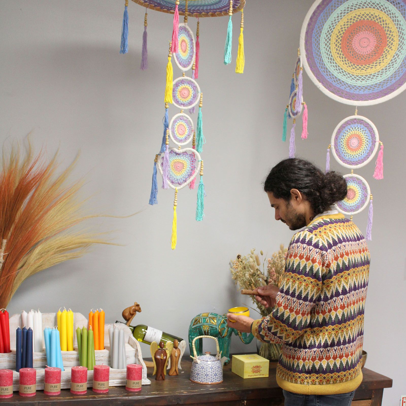 Man holding a small yellow bowl and wooden spoon arranging pottery and colorful candles on a wooden table beneath hanging pastel crocheted dreamcatchers in a boutique-style room Text on candle labels: PURE