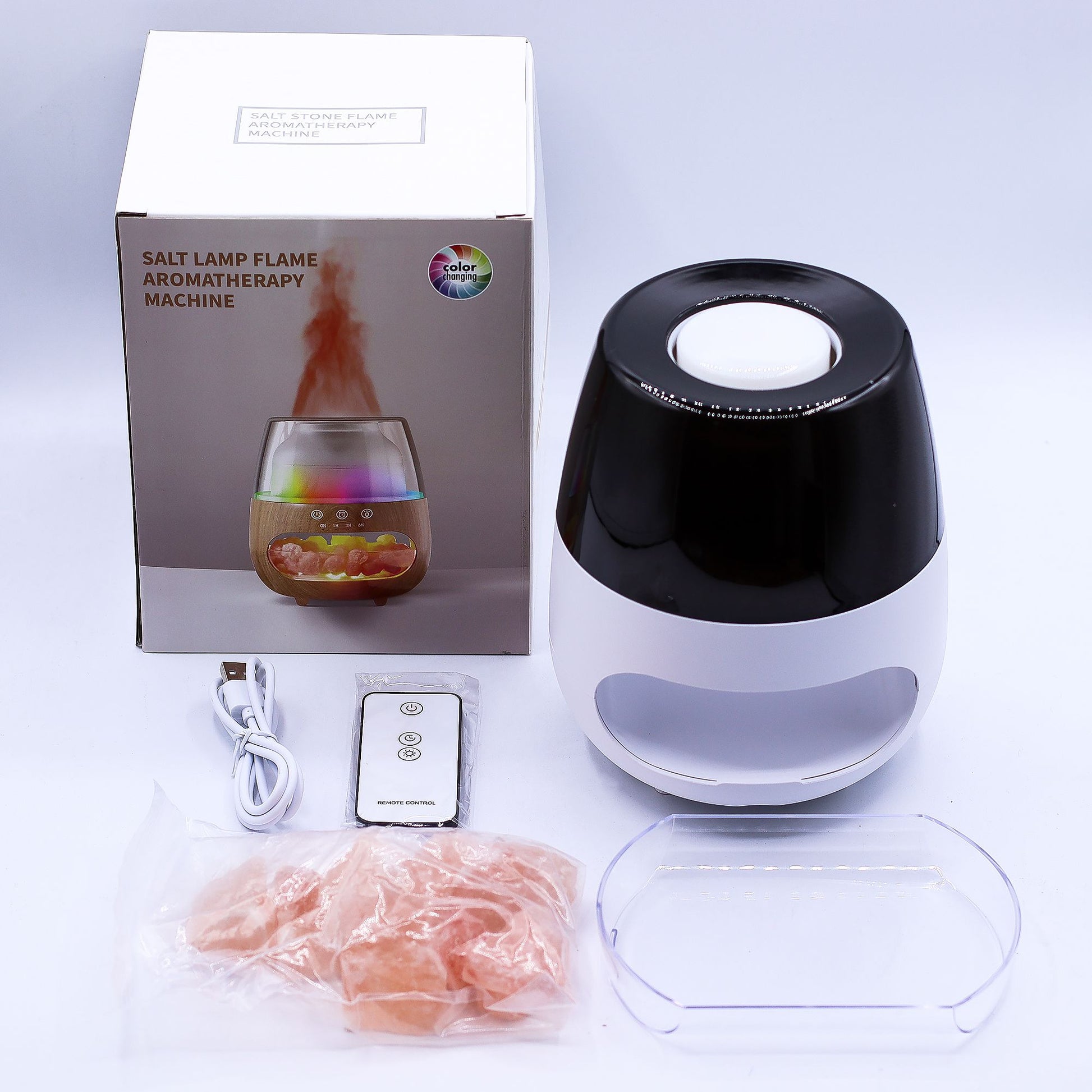 Salt lamp flame aromatherapy machine sits beside its box, USB cable, remote, plastic tray and bag of pink salt on a white surface; box reads "SALT LAMP FLAME AROMATHERAPY MACHINE", top reads "SALT STONE FLAME AROMATHERAPY MACHINE", logo reads "color changing", remote reads "REMOTE CONTROL".