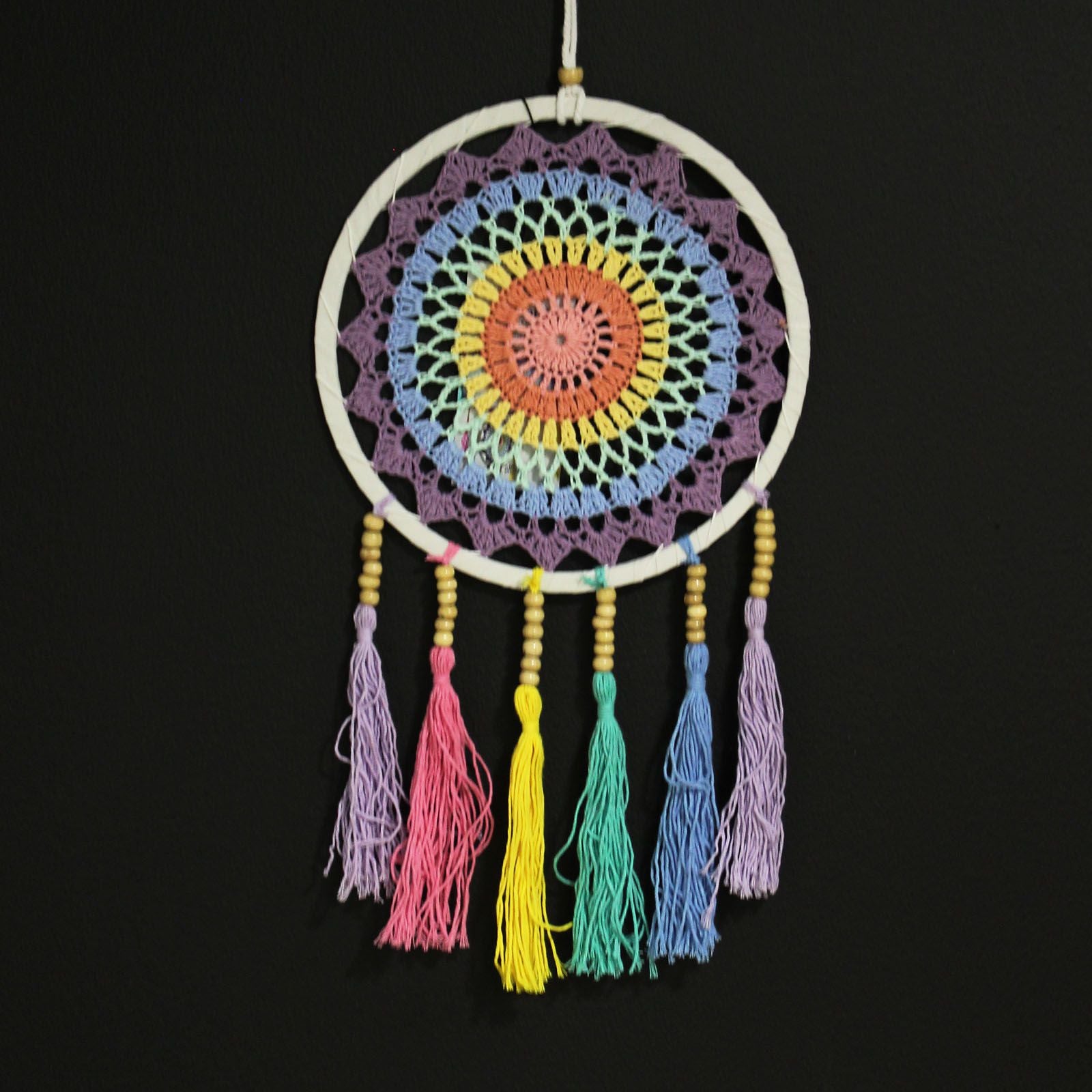 Colorful crocheted circular dreamcatcher hangs from a white cord, showing concentric pastel rings and seven tasseled cords with wooden beads against a plain black wall.