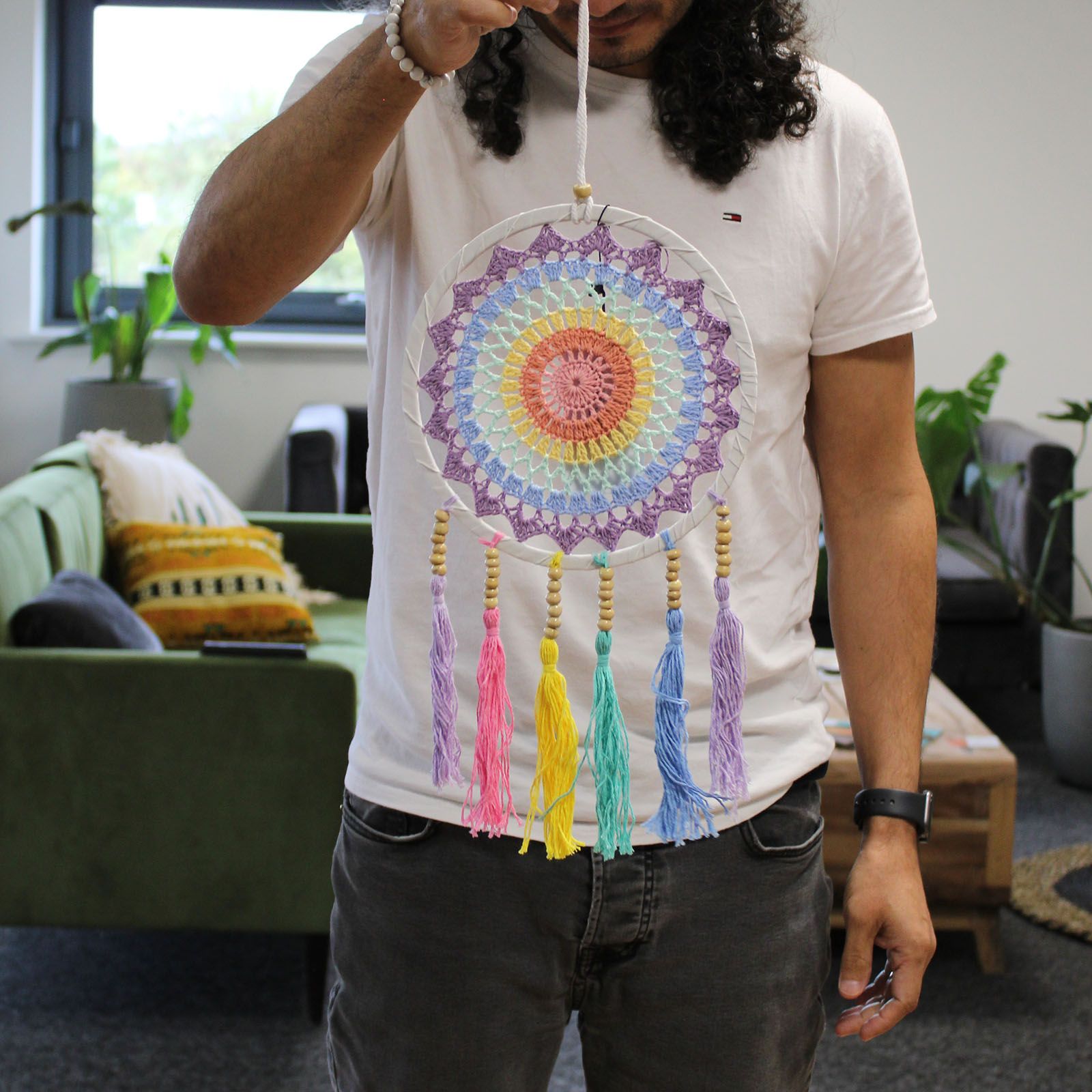 Person holds a colorful crocheted dreamcatcher in front of their chest inside a living room with a green sofa, patterned cushions, plants, and a wooden coffee table.