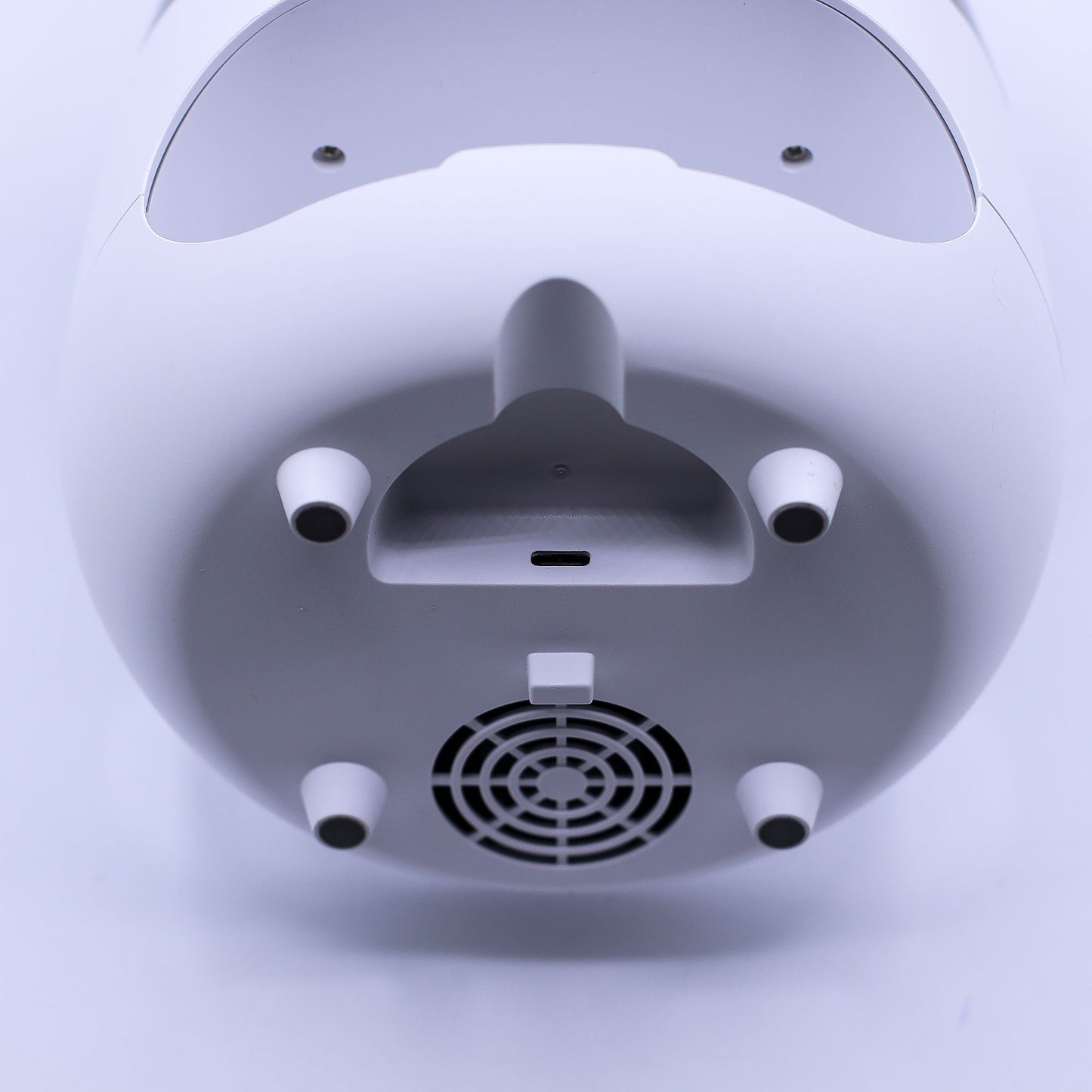 White cylindrical device resting upside down, exposing four conical rubber feet, a central USB-C port and curved handle, a circular ventilation grille, photographed against a plain white background.