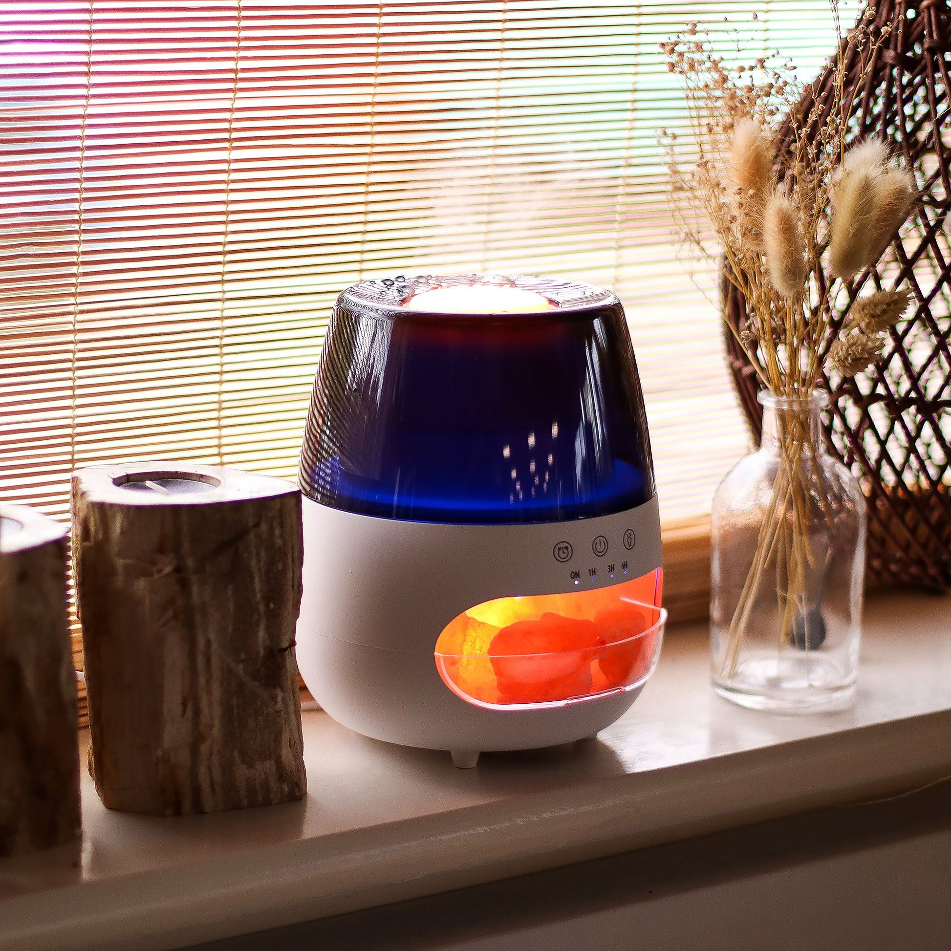 A tabletop diffuser glows orange from visible salt rocks while sitting on a windowsill beside a glass vase of dried grasses and wooden candle holders before bamboo blinds.