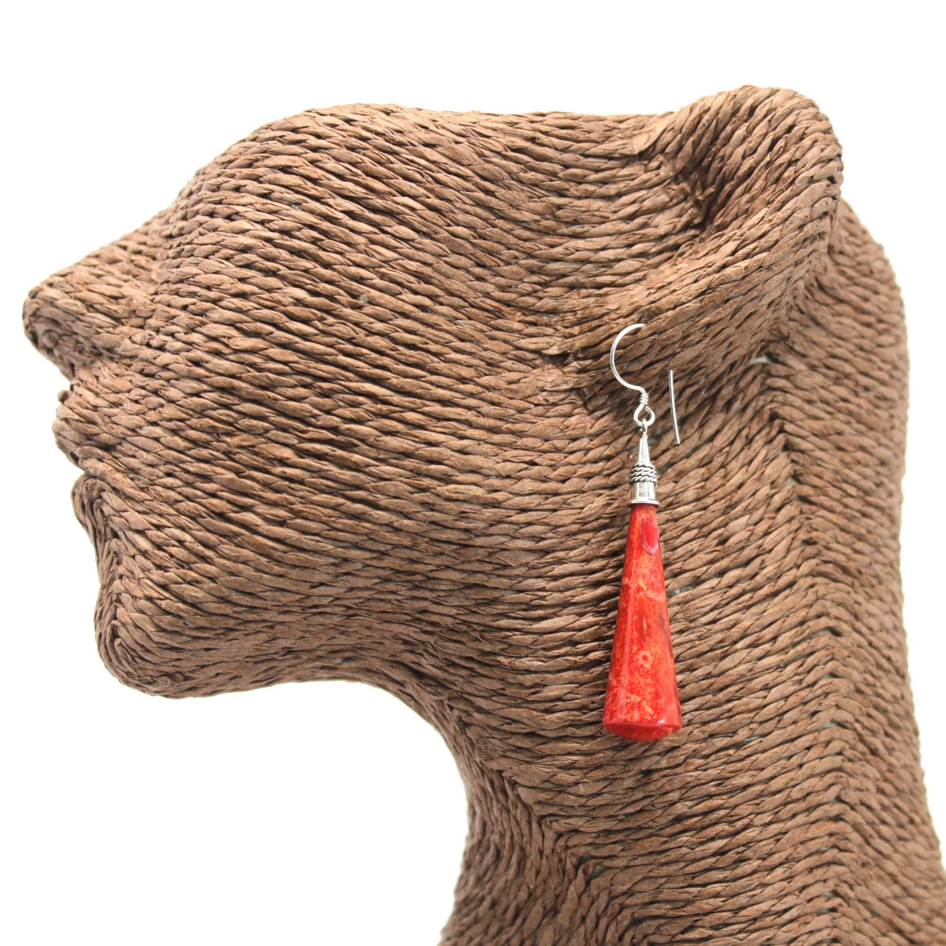 Red cone-shaped dangle earring hangs from the ear of a textured brown woven mannequin head against a white background.
