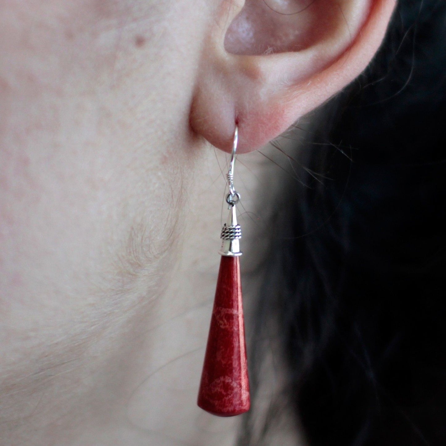 Red cone-shaped dangle earring hangs from a pierced earlobe, silver hook and cap visible, set against dark hair with close-up skin texture.