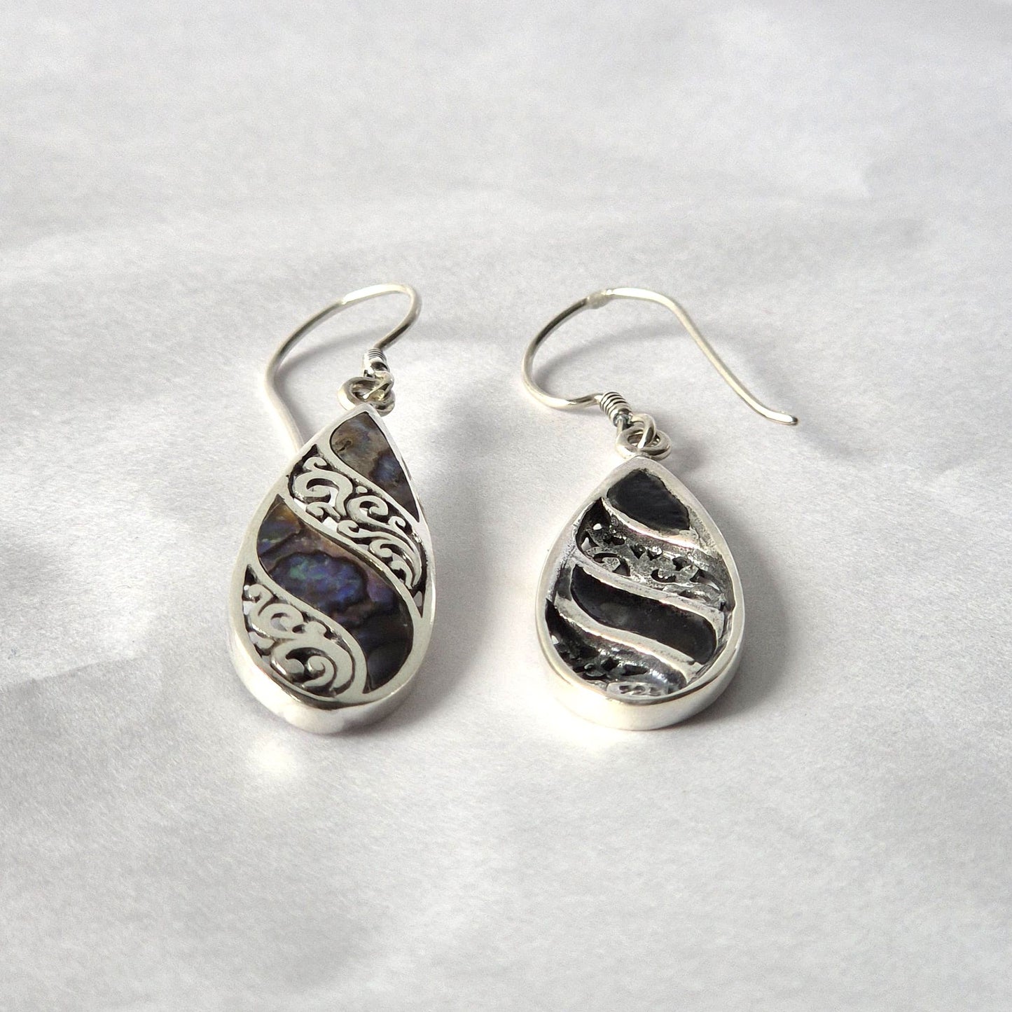 Teardrop silver earrings rest side by side; one features iridescent abalone with ornate scroll filigree, the other darker textured silver; both with fishhook wires on crumpled white paper.