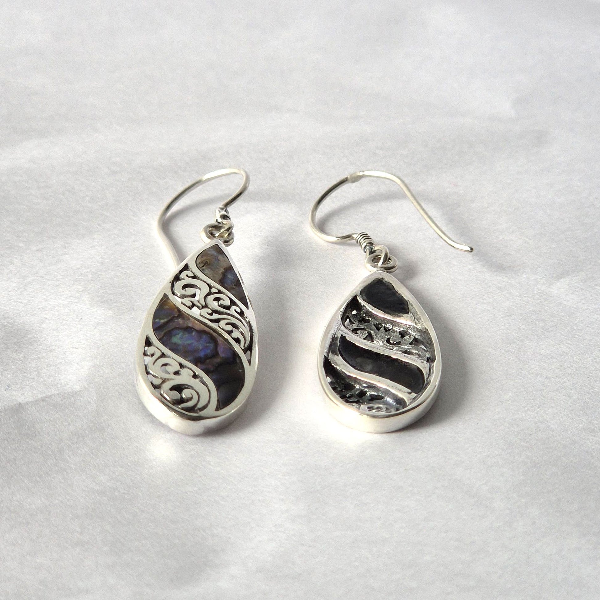 Teardrop silver earrings rest side by side; one features iridescent abalone with ornate scroll filigree, the other darker textured silver; both with fishhook wires on crumpled white paper.