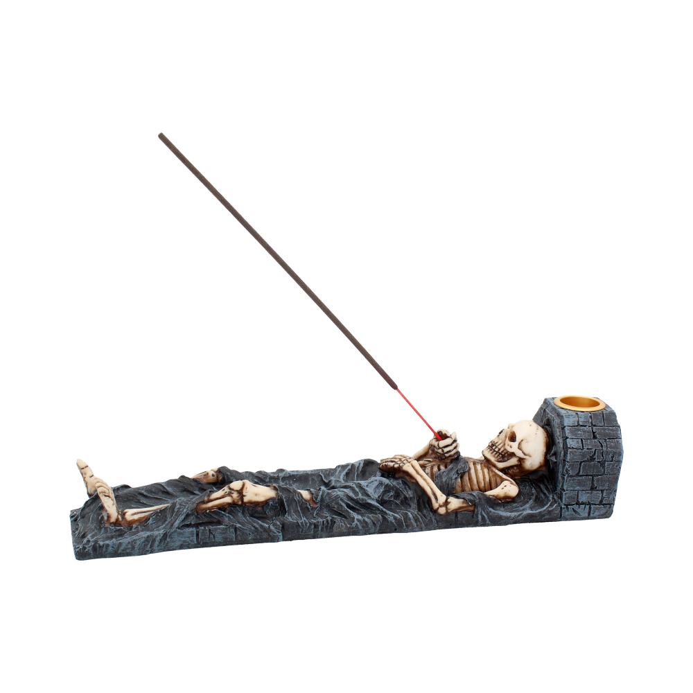 A skeleton figurine holds an incense stick burning over a stone-like base; it is lying on a dark surface.