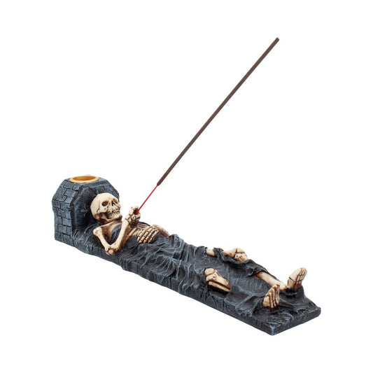 A skeleton figurine lies on a dark incense burner; a lit incense stick rests on its hand.  The piece is isolated on a white background.