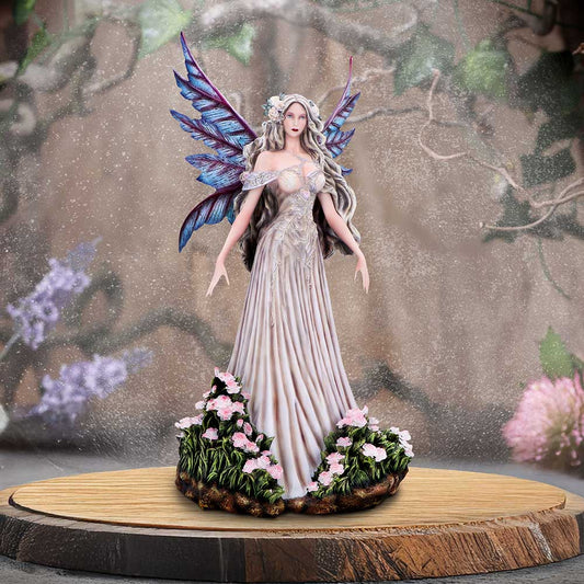 A porcelain fairy figurine stands with outstretched arms on a circular wooden platform, wearing a flowing pale gown, iridescent blue purple wings and floral base amid a blurred garden backdrop.