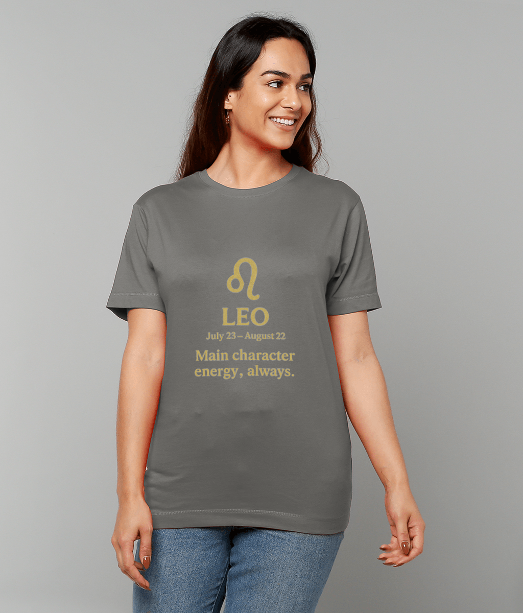 Woman wearing a gray T-shirt displays gold zodiac glyph and text "LEO July 23 - August 22 Main character energy, always." while smiling in front of a plain light-gray studio background.