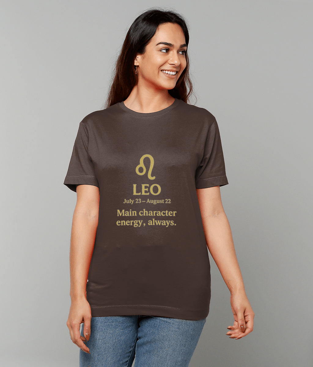 T-shirt displays a gold Leo symbol and the text "LEO July 23 - August 22 Main character energy, always." worn by a smiling woman standing against a plain light-gray studio background.
