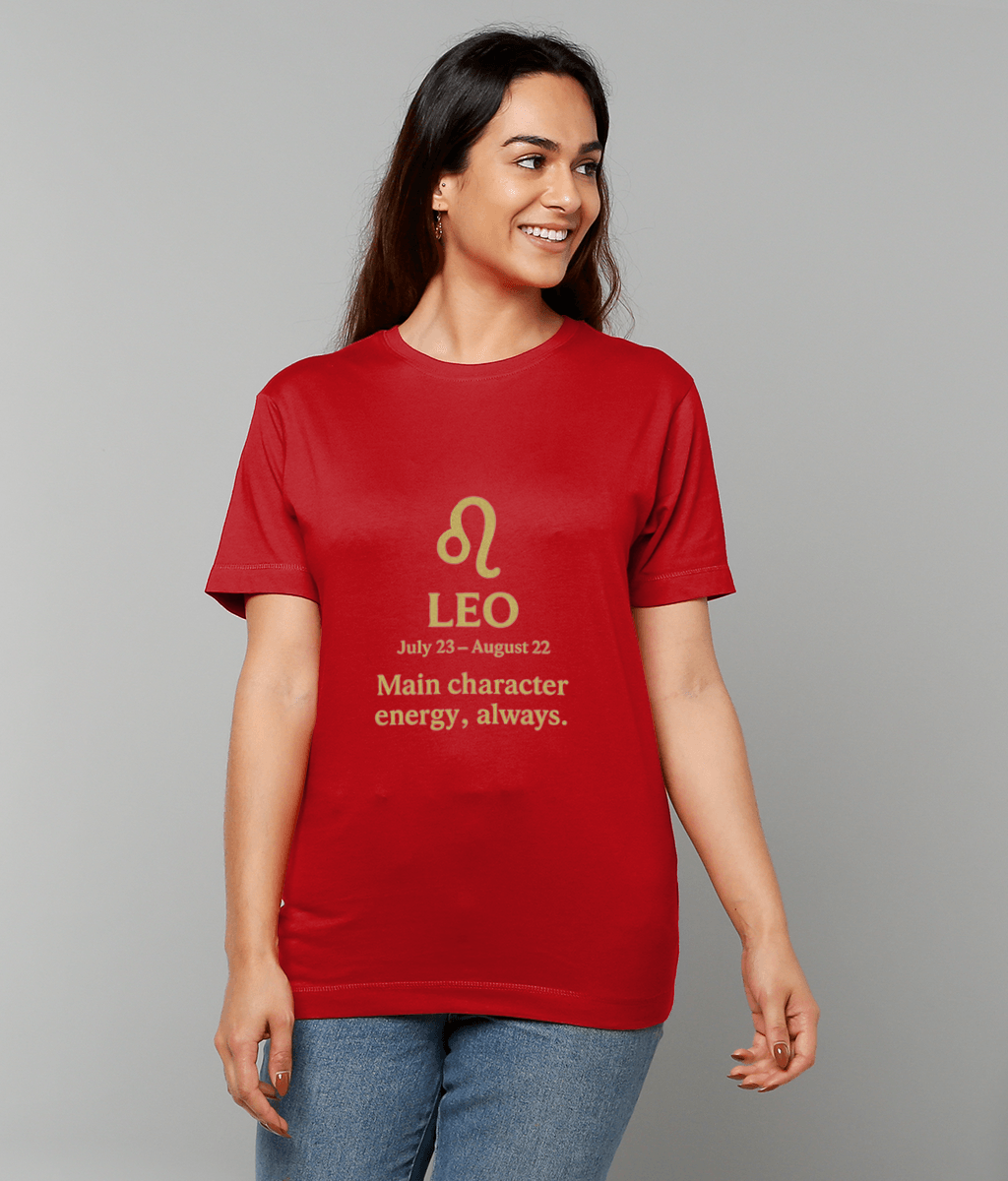 Red T-shirt displaying a gold Leo glyph and text "LEO July 23 – August 22 Main character energy, always." worn by a smiling person standing against a plain gray studio background.