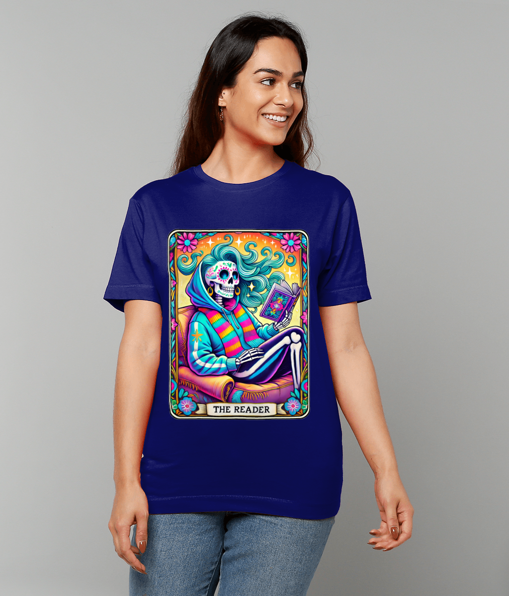 A colorful tarot-style skeleton reads a book, framed by swirling hair and flowers in neon hues on a blue T-shirt worn by a smiling person; text on the design reads "THE READER".
