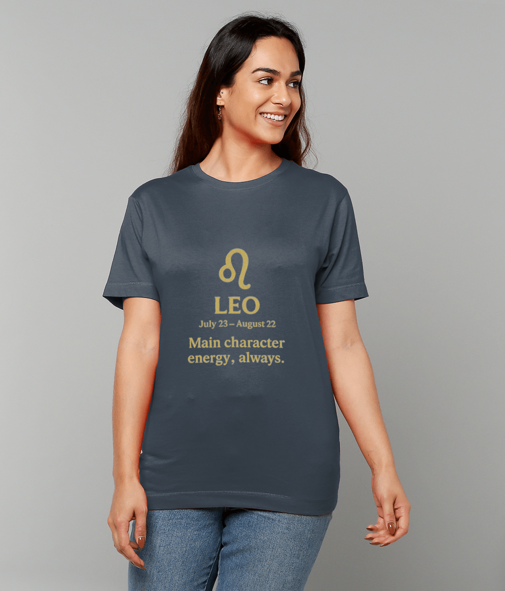 A smiling woman wearing a dark gray t-shirt printed in gold with the Leo symbol and text "LEO July 23 – August 22 Main character energy, always." and jeans, posed against a plain light gray background.