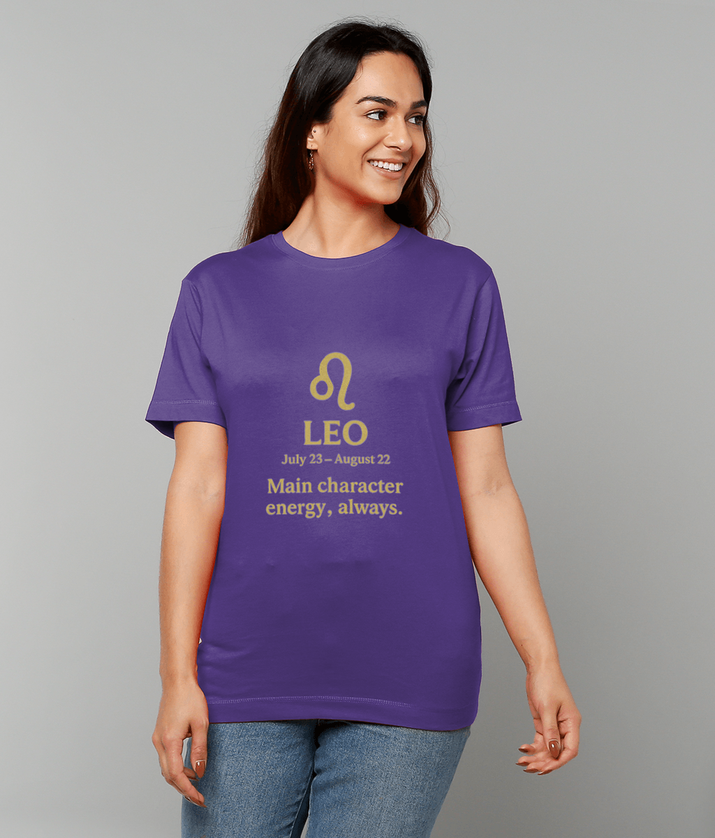 Purple T-shirt with a gold Leo zodiac symbol is worn by a smiling woman looking right against a plain light-gray studio background with jeans; Text on shirt: "LEO July 23 - August 22 Main character energy, always."