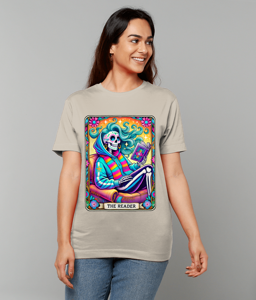 Skull-faced figure reads a book on a colorful tarot-style card labeled "THE READER" while lounging on a chair surrounded by flowers and swirling hair on a beige T-shirt.