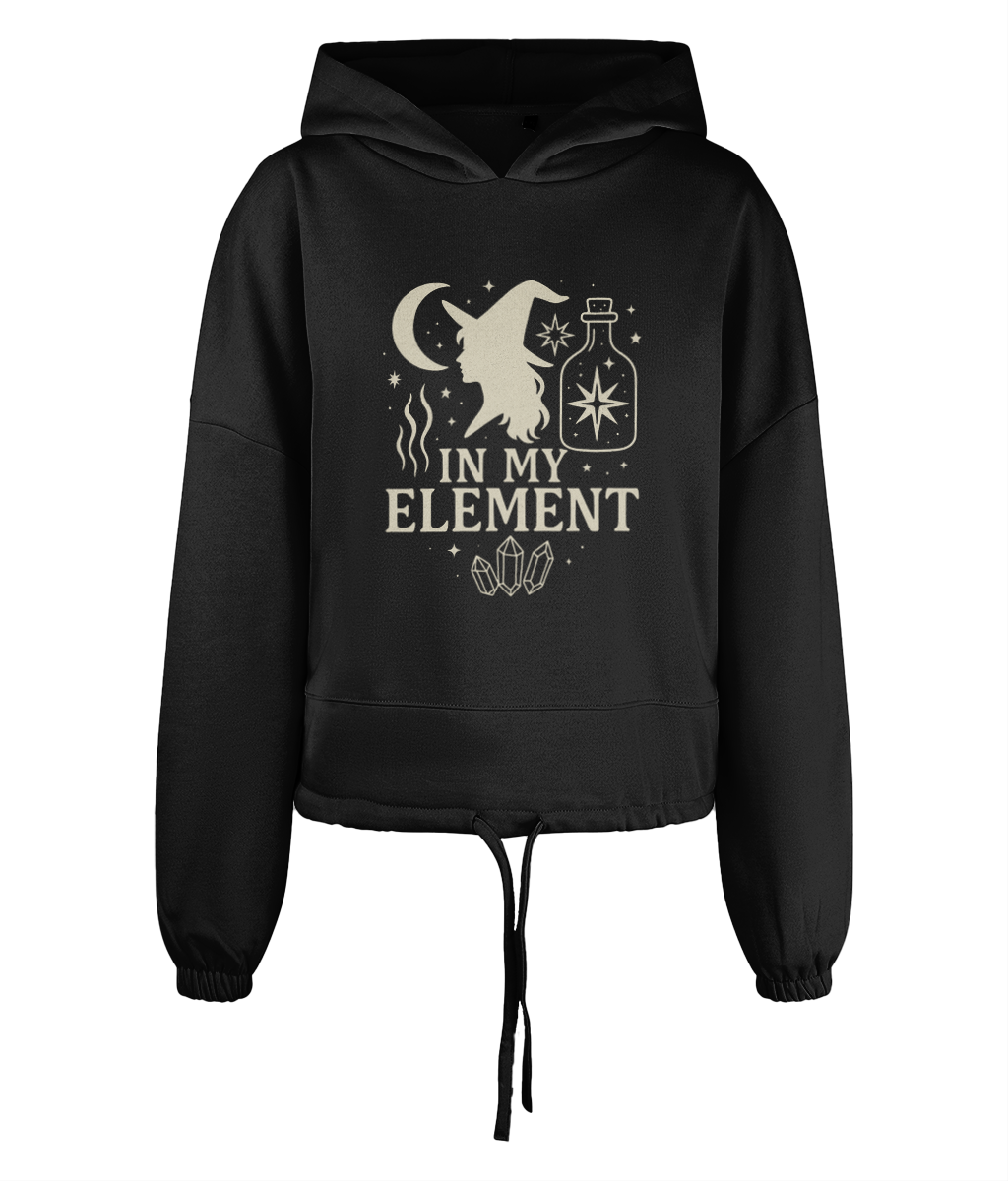Black cropped hoodie displaying a cream witch silhouette, crescent moon, potion bottle, stars and crystals with the text "IN MY ELEMENT" centered on the chest.
