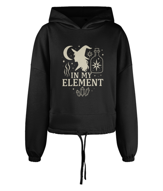 Black cropped hoodie displaying a cream witch silhouette, crescent moon, potion bottle, stars and crystals with the text "IN MY ELEMENT" centered on the chest.