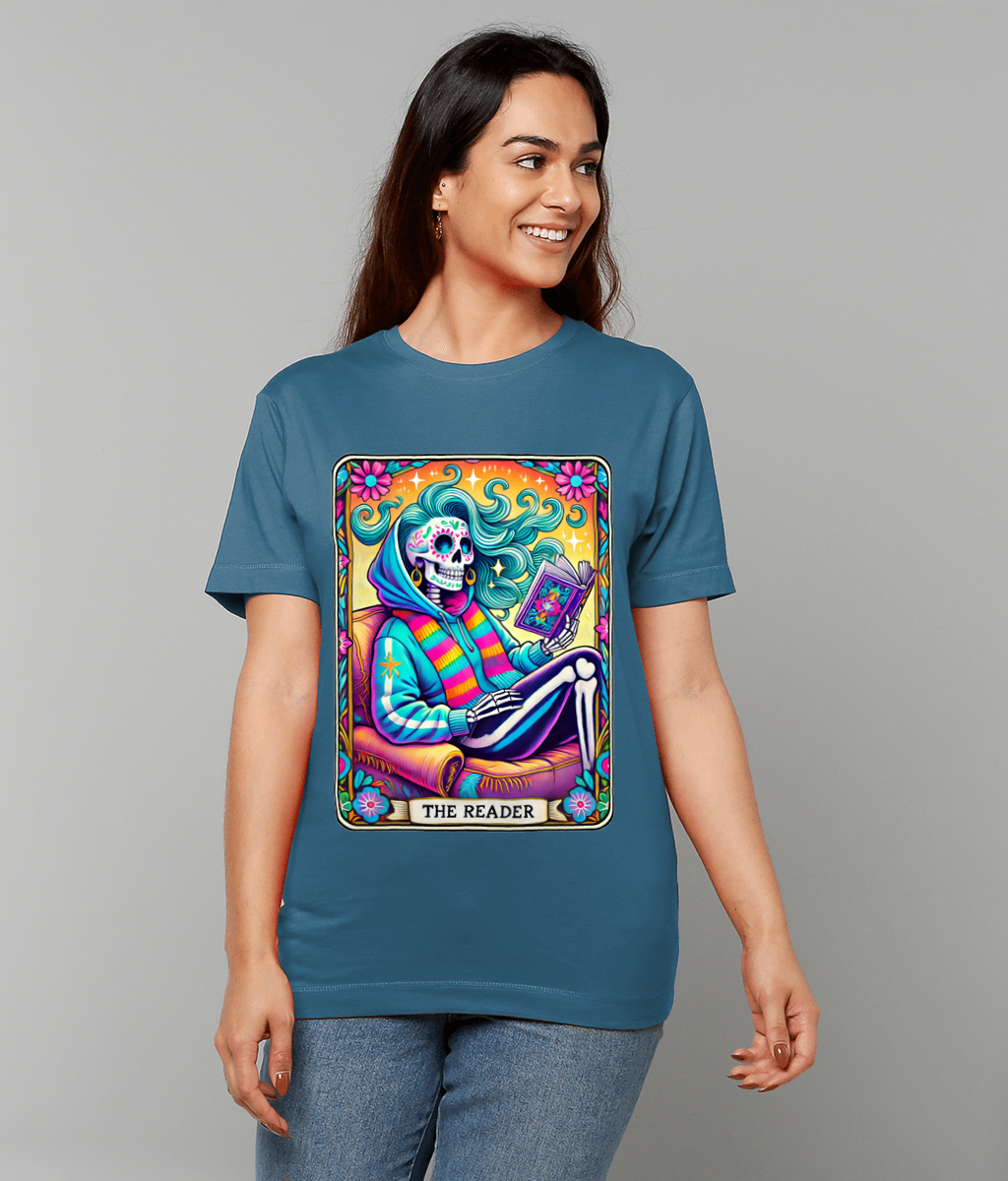 T-shirt shows a colorful tarot-style skeleton wearing a hoodie and striped scarf reading a book while seated in an armchair, decorative floral border, text reads THE READER.