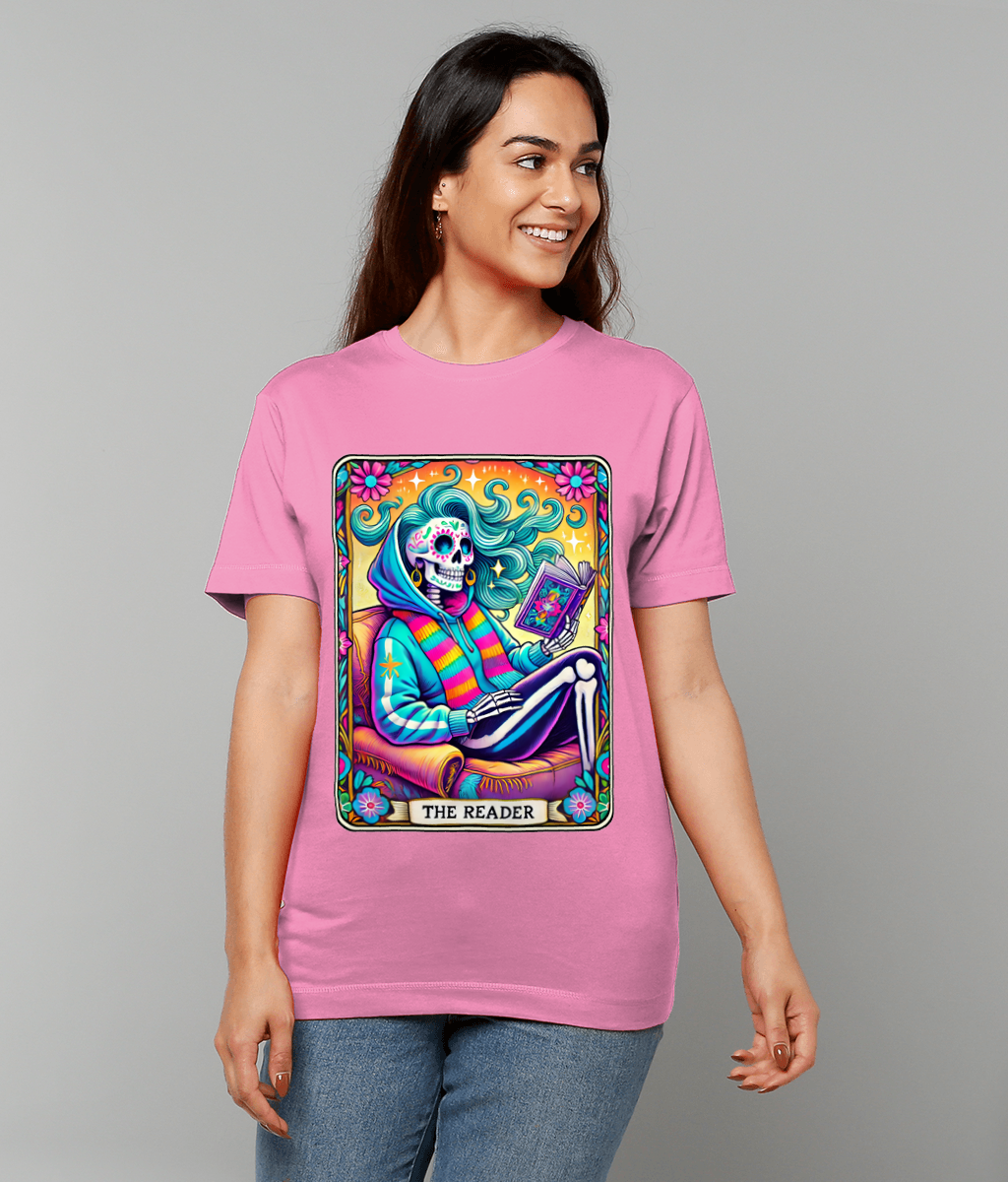 A pink T-shirt displays a colorful skeletal figure reading a book; the caption reads "THE READER"; worn by a smiling woman against a plain gray background.