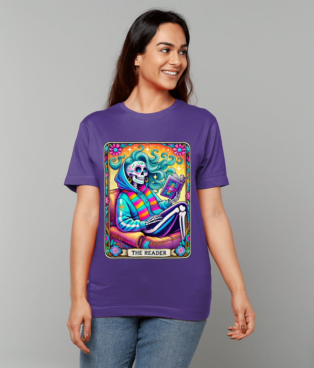 A purple T-shirt displays a colorful tarot-style skeleton reading a book with swirling hair, caption "THE READER", worn by a smiling person against a plain gray background.