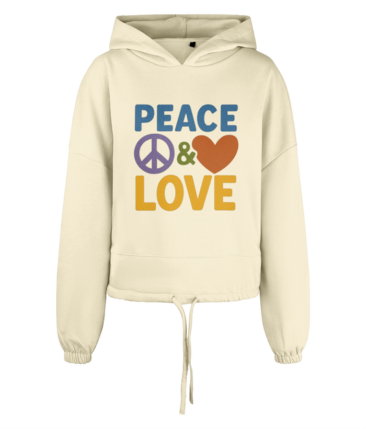 Cream cropped hoodie displays PEACE ☮ & ♥ LOVE printed in bold colorful letters across the front, centered on the garment against a plain black background.