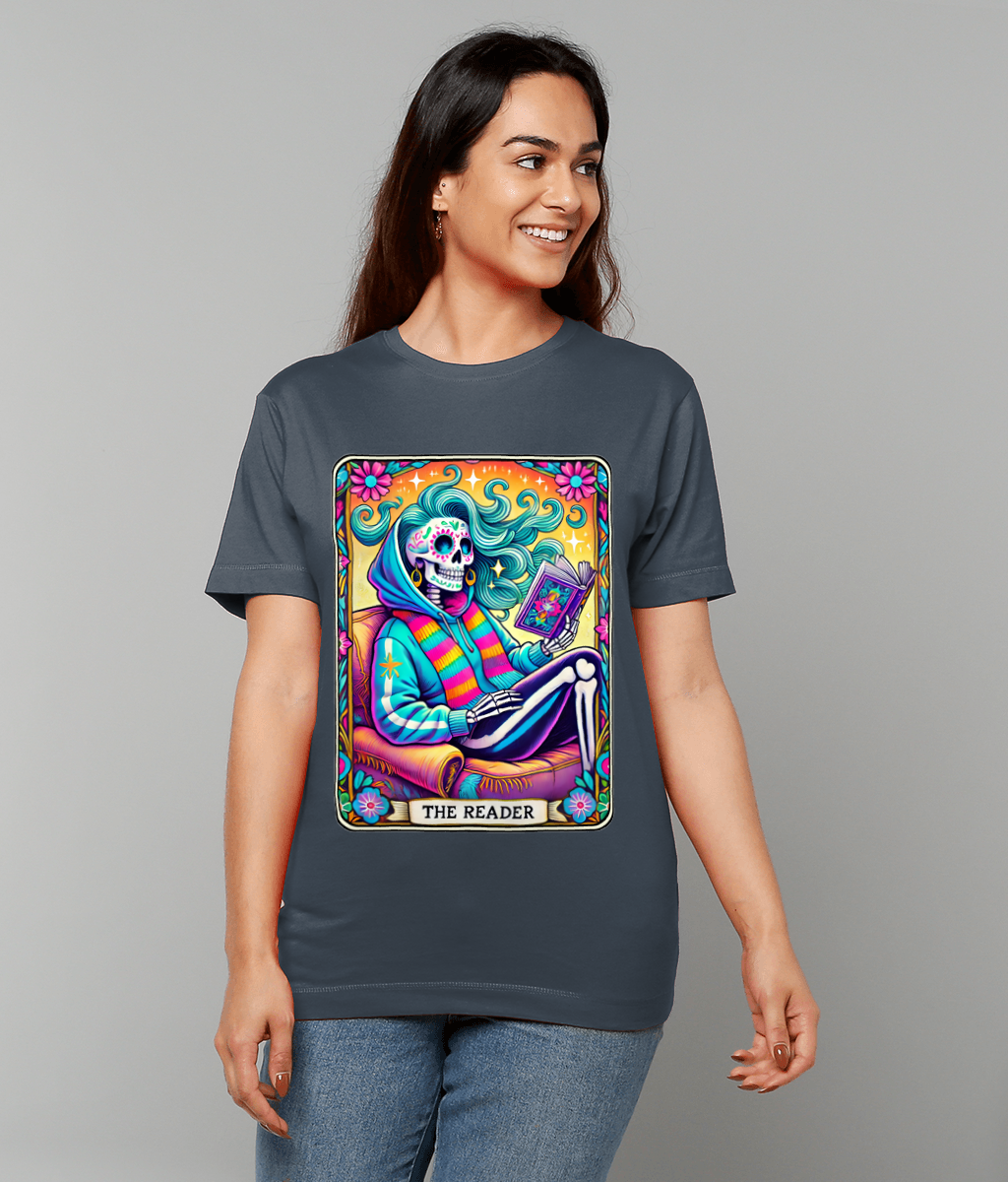 Woman wearing gray T-shirt showing a vibrant tarot-style skeleton in a hoodie reading a book while seated in an armchair, surrounded by flowers and stars, caption "THE READER" on design.