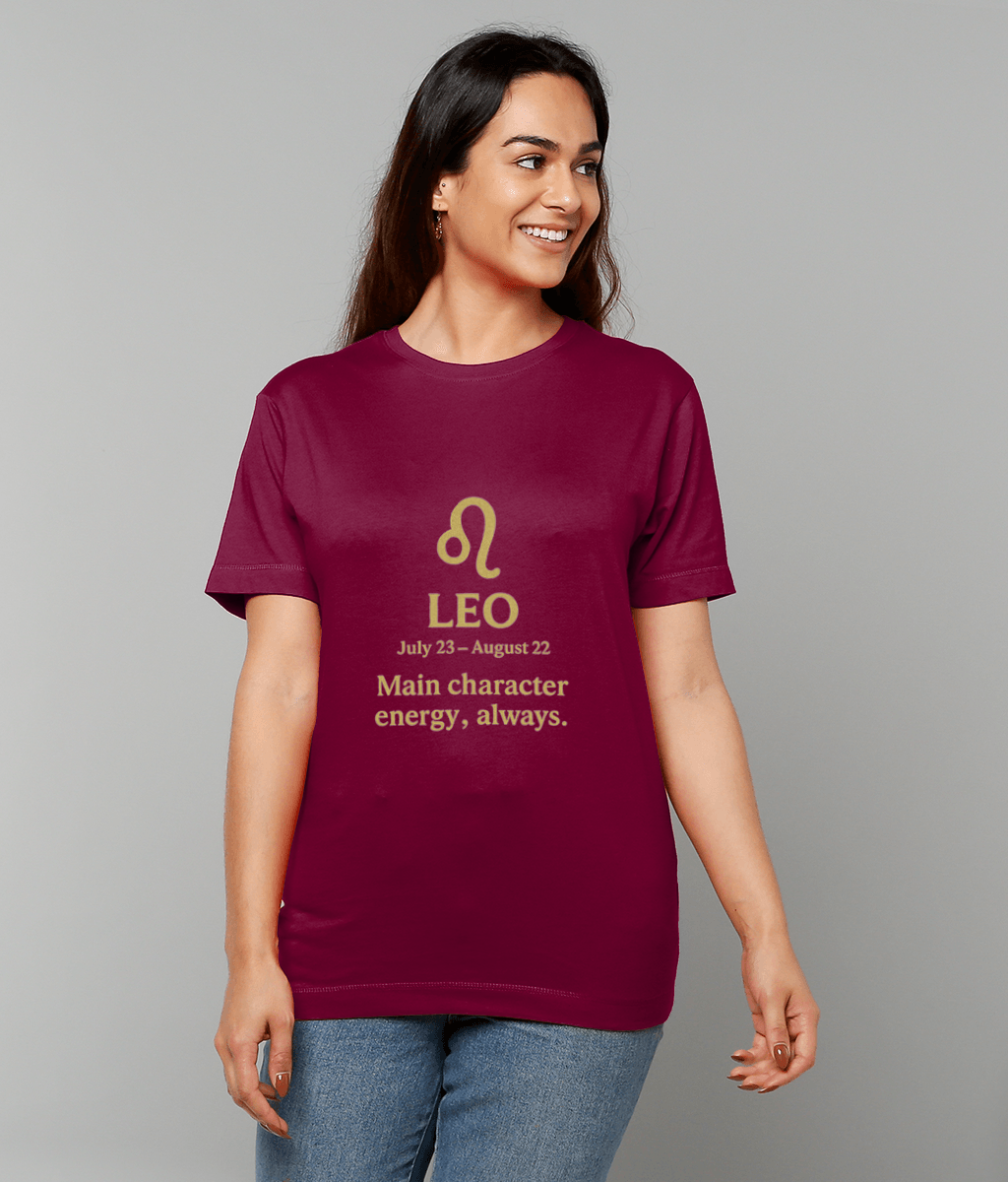 Woman wearing a maroon T-shirt with a gold Leo symbol, smiling and looking to her right against a plain gray backdrop while wearing jeans; T-shirt text: LEO July 23 – August 22 Main character energy, always.