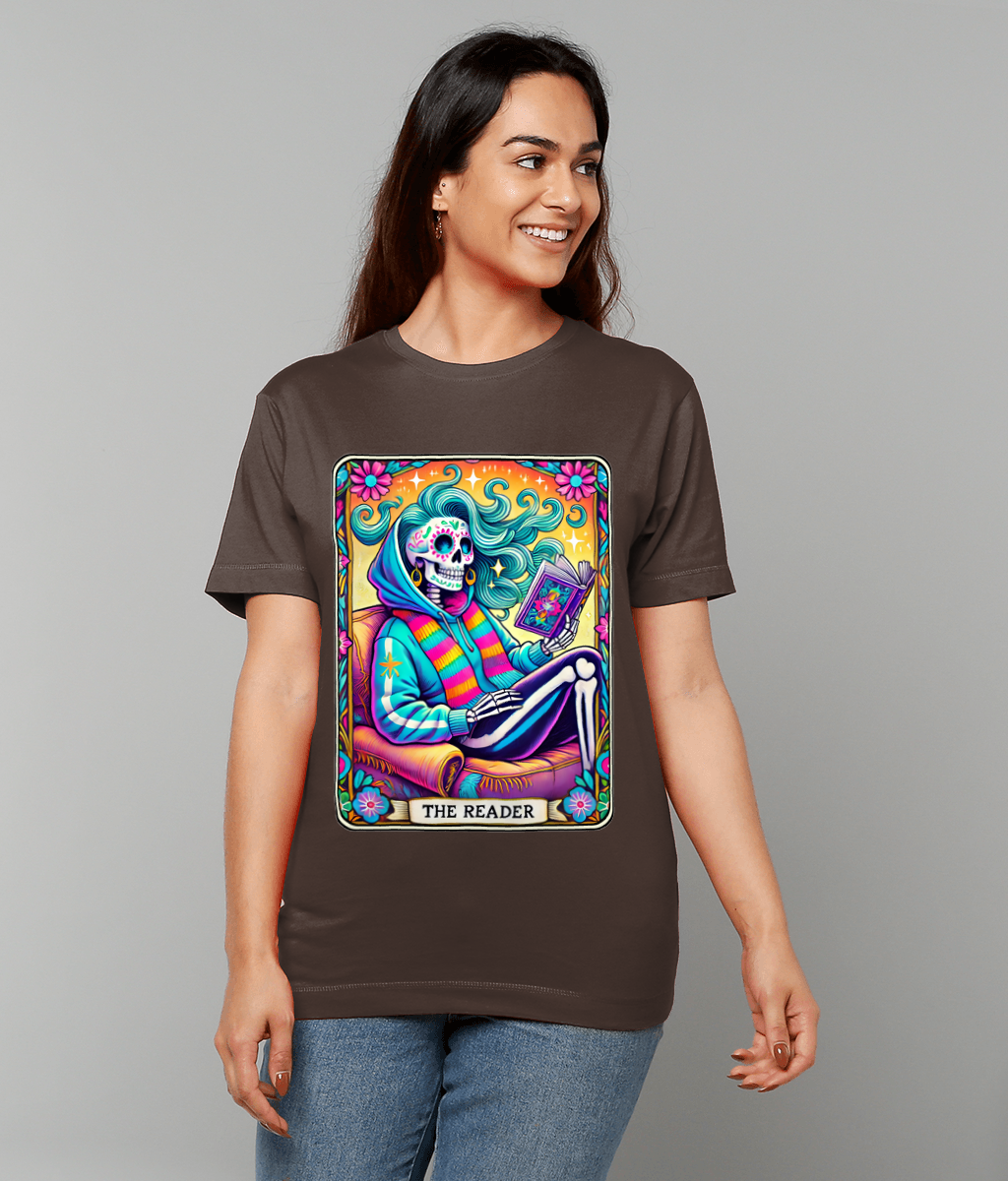 Woman wearing brown t-shirt displaying a colorful tarot-style illustration of a skeleton reading a book while seated in an armchair surrounded by flowers, text: THE READER.