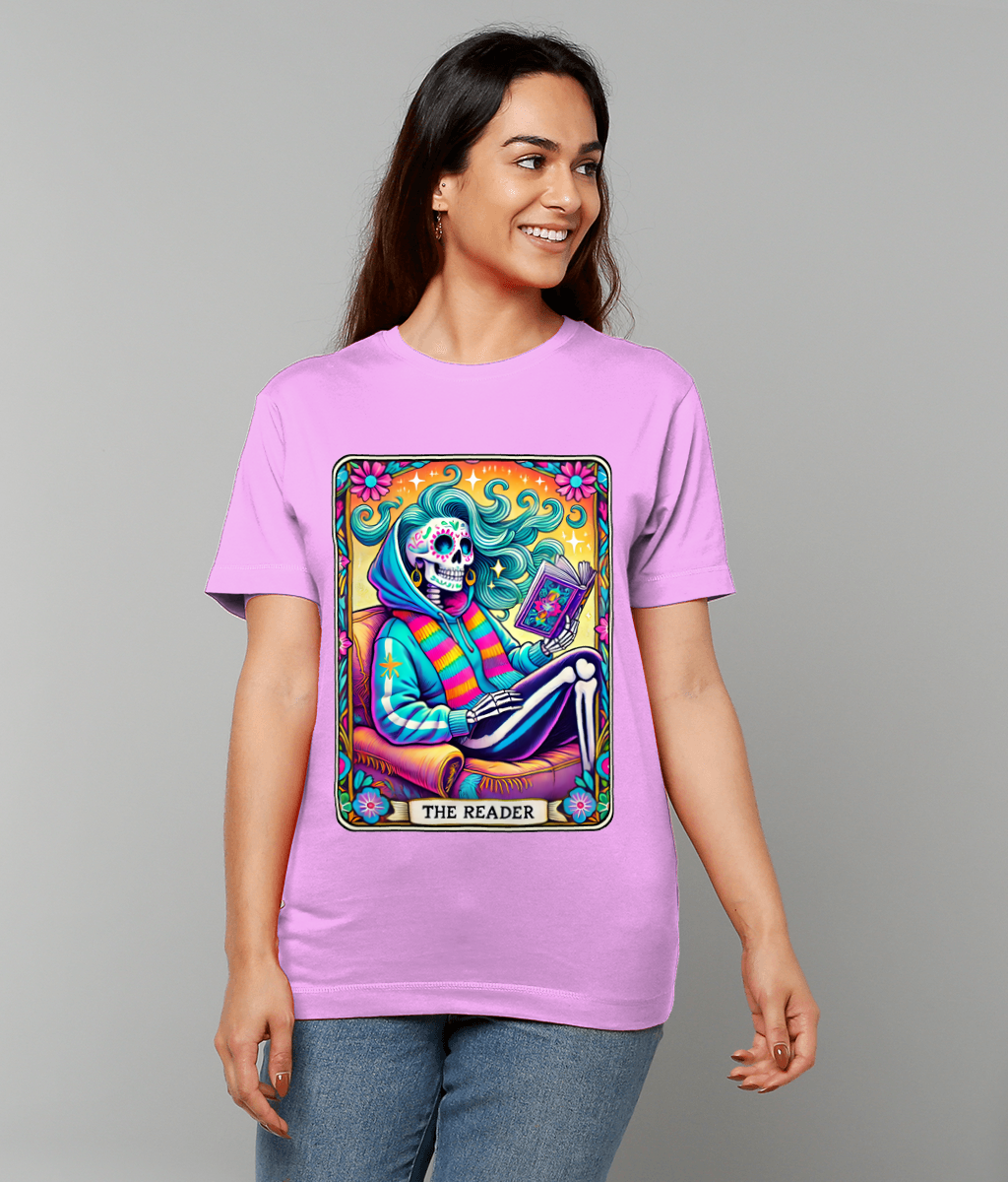 A vibrant skeleton reads a book while reclining in an armchair, printed on a lavender T-shirt worn by a smiling woman against a plain gray background Text: THE READER