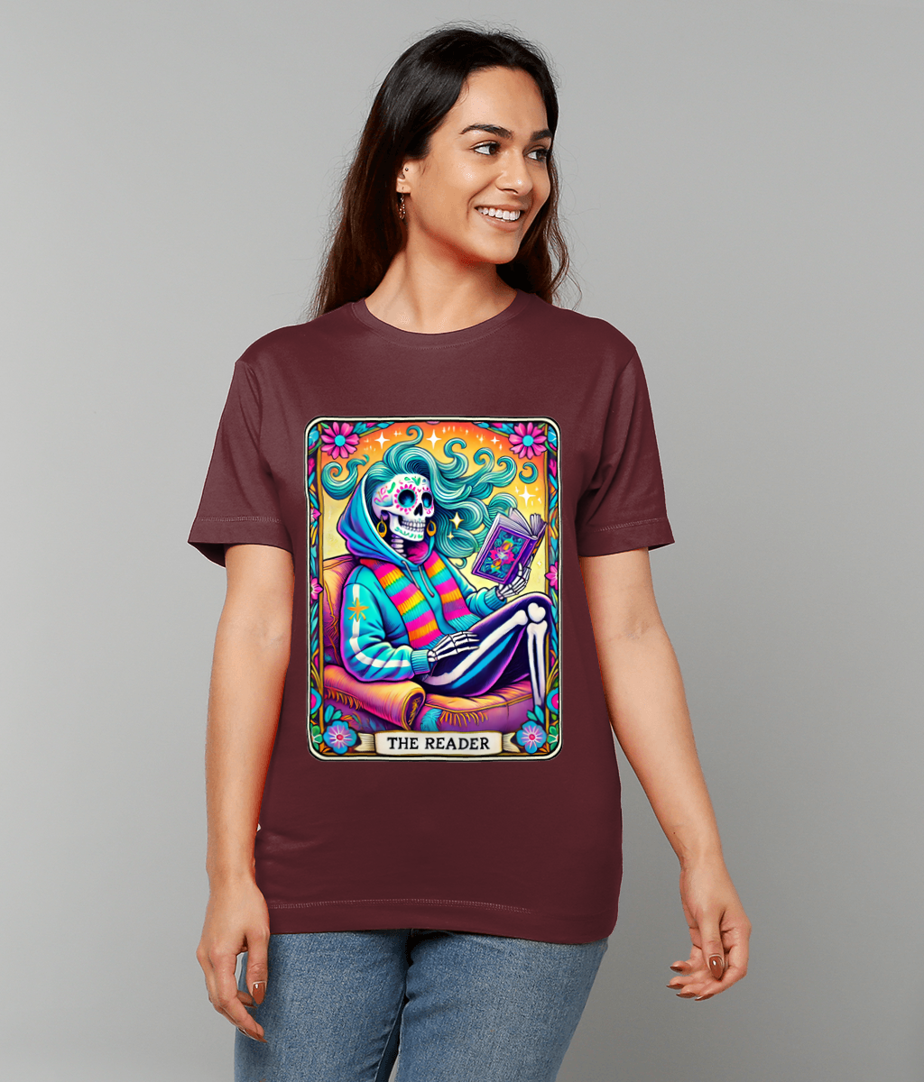 Colorful sugar-skull skeleton sits reading a book on a vibrant tarot-like card printed on a maroon T-shirt worn by a smiling woman against a plain gray background, text: THE READER