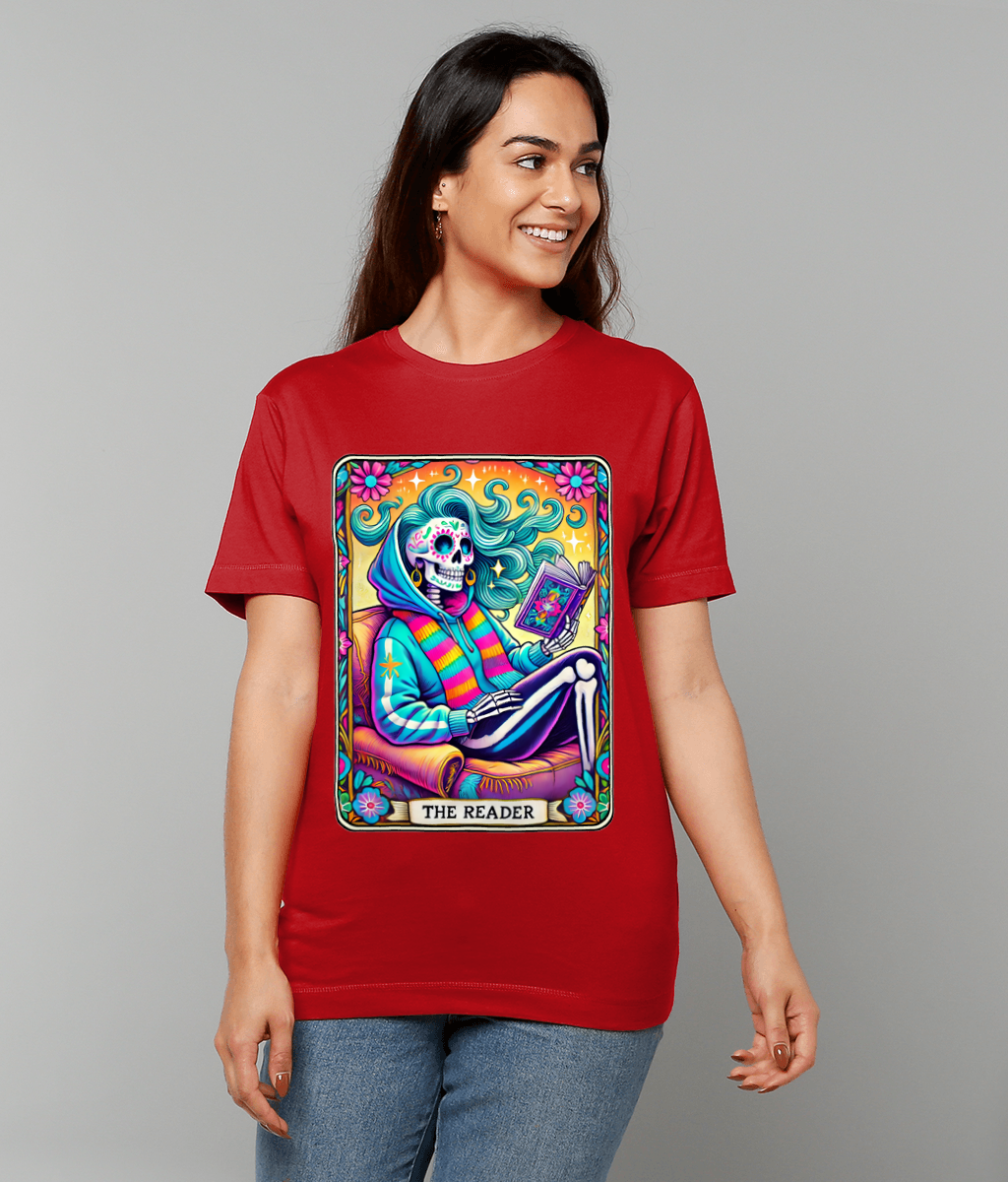 Woman in red T-shirt shows a colorful tarot-style illustration of a smiling skeleton reading a book, seated in an armchair; design caption reads 'THE READER' and background is plain gray.