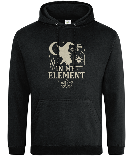 Black hoodie displays cream graphic of a witch silhouette, crescent moon, potion bottle, crystals and stars with the text 'IN MY ELEMENT' centered on the chest against a plain background.