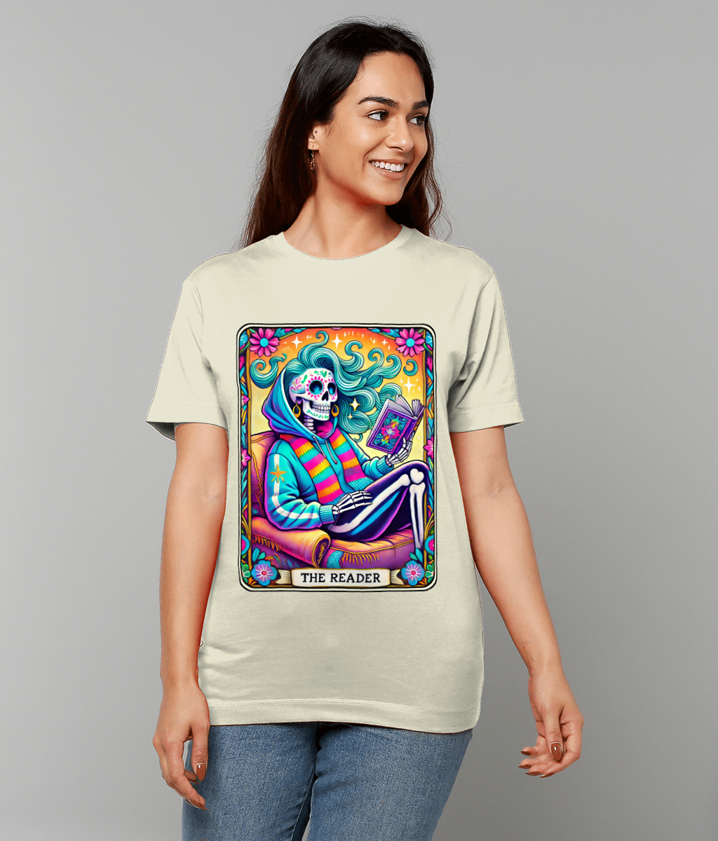 A colorful skeletal figure lounges reading a book within a vibrant floral tarot-style panel printed on a beige T-shirt; caption at bottom reads THE READER.