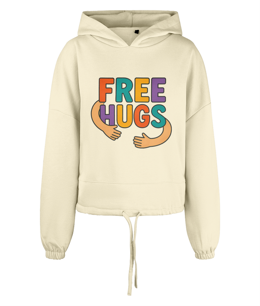 Cream cropped hoodie displays large colorful graphic reading "FREE HUGS" with cartoon arms forming a hug across the chest, shown centered against a plain black background.