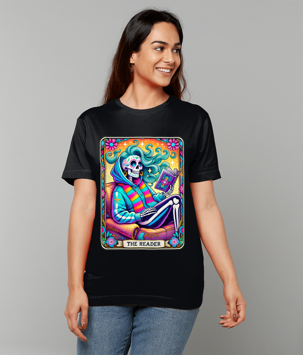 A vibrant tarot-style skeleton sits reading a book on a black T-shirt worn by a smiling woman against a plain gray background, design text: THE READER