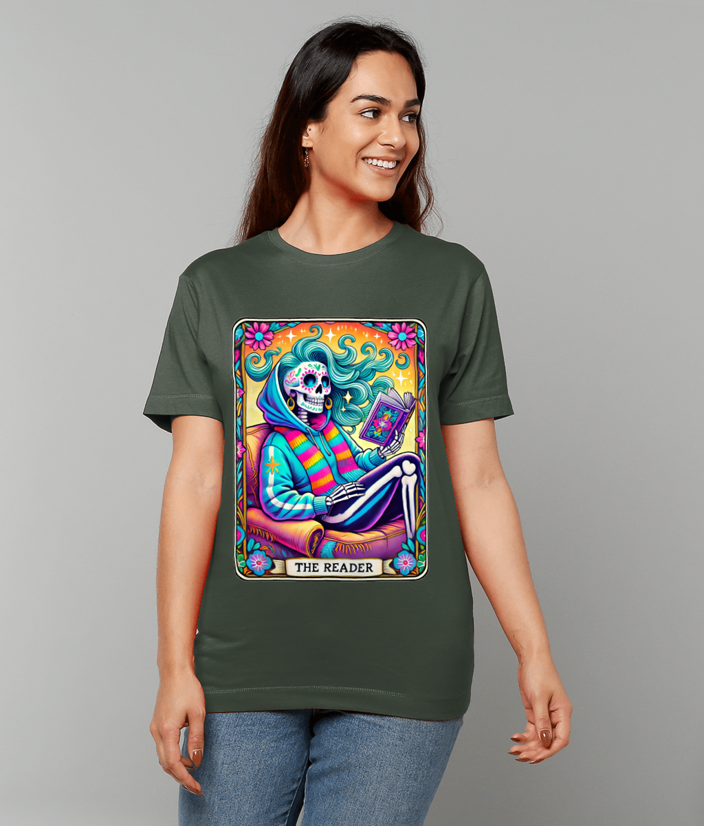 Colorful skeletal figure reads a book while reclining in a cushioned chair, framed by a vivid floral border on an olive T-shirt worn by a smiling person in a neutral studio; text: THE READER.