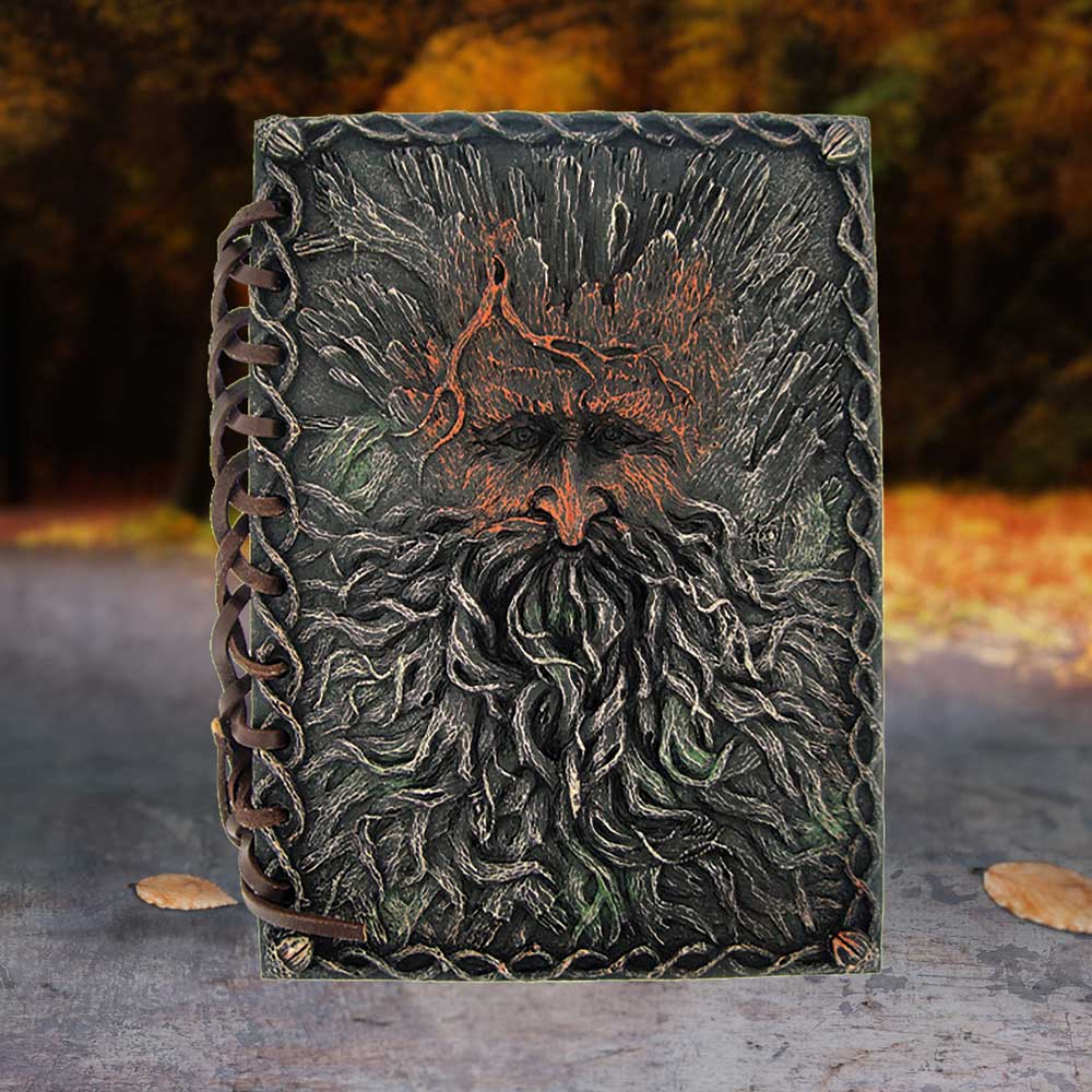 A detailed leather journal features an embossed face with a long beard, surrounded by a braided border. It lies on a stone surface with a blurred, autumnal forest backdrop.